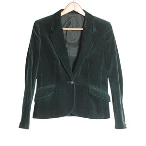 May include: A dark forest green corduroy blazer featuring a single-button closure, notched lapels, and two front flap pockets. The jacket is fully lined with a dark interior fabric and is displayed on a wooden hanger against a plain background.