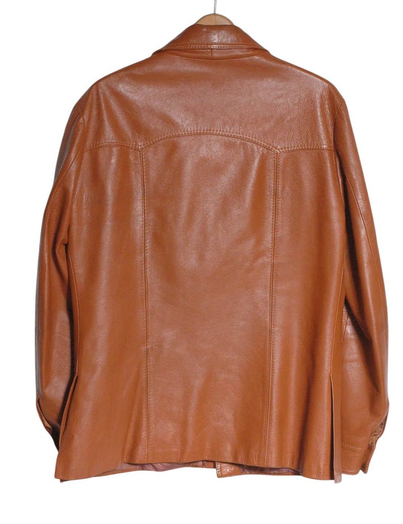 May include: A brown leather jacket hanging on a wooden hanger. The jacket has a classic design with a collar, button closure, and pockets. The leather appears to be smooth and has a slight sheen. The jacket is a medium brown color.