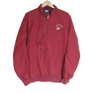 Peut inclure: Un coupe-vent Nike bordeaux à enfiler avec une fermeture éclair à mi-hauteur, un col montant et une grande poche ventrale. La poitrine présente une broderie blanche avec le texte Athletics West au-dessus du logo et du swoosh Nike.