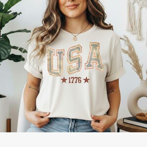 Könnte beinhalten: Ein cremefarbenes Kurzarm-T-Shirt mit dem Schriftzug USA in großen, abgenutzten Blockbuchstaben in einem gedämpften Blau- und Orangeton. Unter dem Text ist die Zahl 1776 von zwei kleinen roten Sternen im passenden Used-Look umrahmt.