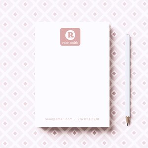 May include: A white notepad with a pink logo featuring the letter "R" and the text "rose smith". The notepad has a pink and white geometric pattern background. The text "rose@email.com - 987.654.3210" is printed at the bottom of the notepad.