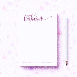 May include: A white notepad with a pink floral pattern background. The notepad has a pink cursive name "Katherine" at the top and the text "katherine@email.com 765 432 1098" at the bottom. A white pencil is lying next to the notepad.