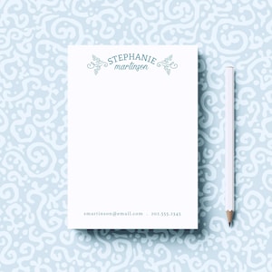 May include: A white notepad with a blue floral design and the text "STEPHANIE martinson" at the top. The notepad has a white pencil next to it and the text "smartinson@email.com 202.555.2345" at the bottom.