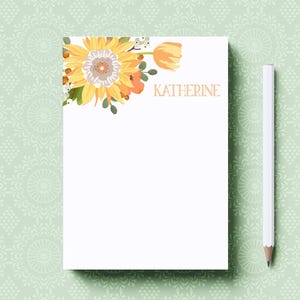 May include: White notepad with floral sunflower and tulip design. The name "KATHERINE" is printed in gold. A white pencil is placed to the right of the notepad. The background is a light green floral pattern.