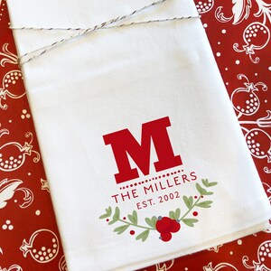 May include: White kitchen towel with a red initial "M" and the text "THE MILLERS EST. 2002". The design includes a green and red berry wreath. The towel is tied with twine.