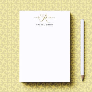 May include: A white notepad with a gold monogram of the letter "R" and the name "Rachel Smith" printed at the top. The notepad is on a yellow background with a swirling pattern.