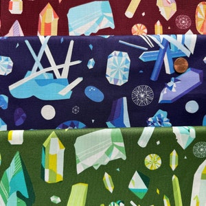 Może przedstawiać: Trzy próbki tkaniny z różnymi geometrycznymi wzorami kolorowych kryształów. Górna próbka jest bordowa z różowymi, żółtymi i białymi kryształami. Środkowa próbka jest granatowa z niebieskimi, białymi i fioletowymi kryształami. Dolna próbka jest zielona z zielonymi, żółtymi i białymi kryształami.