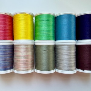 May include: Ten spools of thread arranged in two rows of five. The top row features solid colors in red, yellow, bright green, blue, and dark purple. The bottom row displays variegated or metallic-toned threads in blue, beige, olive, silver, and brown.