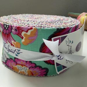 Może przedstawiać: Rolka materiału z kwiatowym wzorem w odcieniach różu, pomarańczy i turkusu. Materiał jest owinięty białą wstążką z napisem "Free Spirit". Widoczne są krawędzie materiału, ukazujące różnorodność kolorów.