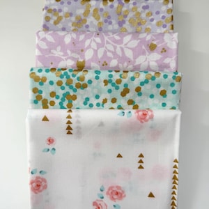 Może przedstawiać: Stos złożonych tkanin z różnymi wzorami kwiatowymi i geometrycznymi. Tkaniny mają kolory różowy, fioletowy i turkusowy ze złotymi akcentami. Dolna tkanina ma różowe róże i złote trójkąty.