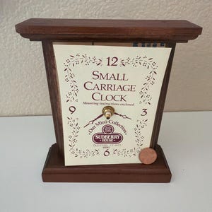 Puede incluir: Un pequeño reloj de viaje de madera con acabado marrón oscuro. La esfera del reloj es blanca con números negros y el texto "SMALL CARRIAGE CLOCK". Una moneda de cobre descansa sobre la base. El reloj es de la "Mini-Collection".