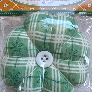 Może przedstawiać: Zielony i biały w kratę poduszeczka na szpilki do nadgarstka w kształcie kwiatu z białym guzikiem w środku. Etykieta głosi "Wrist Pin Cushion by Lori Holt of Bee in my Bonnet".