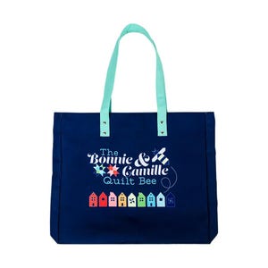 Puede incluir: Una bolsa de lona azul con el texto "The Bonnie & Camille Quilt Bee" y un diseño de abeja y casas de colores.