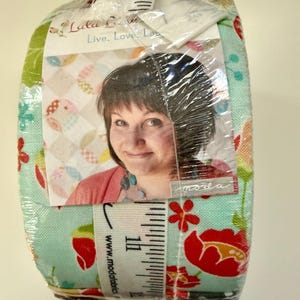 Może przedstawiać: Pakiet tkanin owinięty w przezroczysty plastik. Tkanina ma kwiatowy wzór z czerwonymi kwiatami na turkusowym tle. Na przodzie widoczne jest zdjęcie kobiety wraz z tekstem "Lulu Lane" i "Live. Love. Laugh."