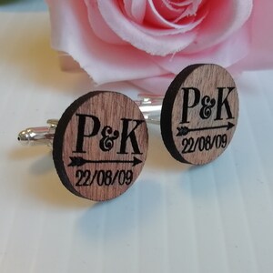 Puede incluir: Un par de gemelos de madera con respaldo plateado. Los gemelos son redondos y tienen una flecha negra apuntando hacia la derecha con las iniciales "P&K" y la fecha "22/08/09" grabadas en ellos.