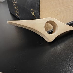 May include: A wooden book page holder with a rounded opening in the center. The holder is light brown and has a smooth finish.
