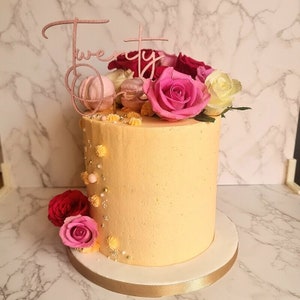 May include: A yellow cake decorated with pink and white macarons, pink and white roses, and a pink "Twenty Five" cake topper. The cake is on a white cake stand with a gold ribbon.