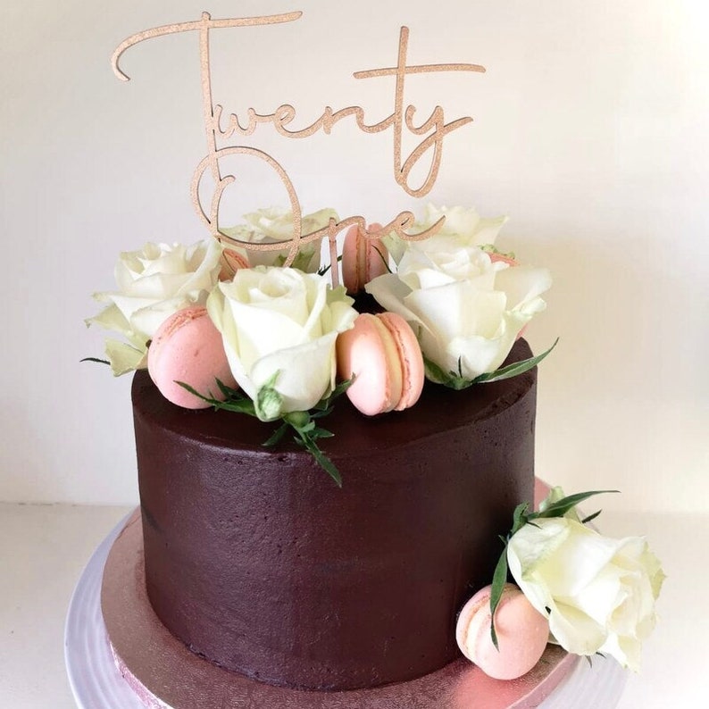 May include: A chocolate cake decorated with white roses and pink macarons. A wooden cake topper says "Twenty One" in cursive.