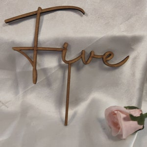 Puede incluir: Adorno de pastel de madera marrón con la palabra "Five" escrita en cursiva. El adorno tiene un palito para insertar en el pastel. Una rosa rosa se encuentra en la esquina inferior derecha.