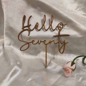 Puede incluir: Topper de pastel de madera con el texto "Hello Seventy" en una fuente cursiva. El topper es marrón y tiene un palo unido a la parte inferior para insertar en un pastel.