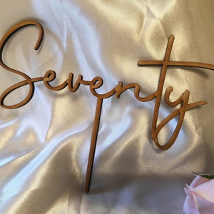 Puede incluir: Topper de pastel de madera con la palabra "Setenta" escrita en cursiva. El topper es marrón y está sobre un fondo blanco.