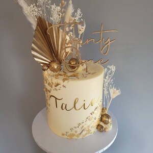 May include: A white cake decorated with gold accents, dried flowers, and a wooden topper that says "Twenty One". The cake is also decorated with the name "Talia" in gold lettering.