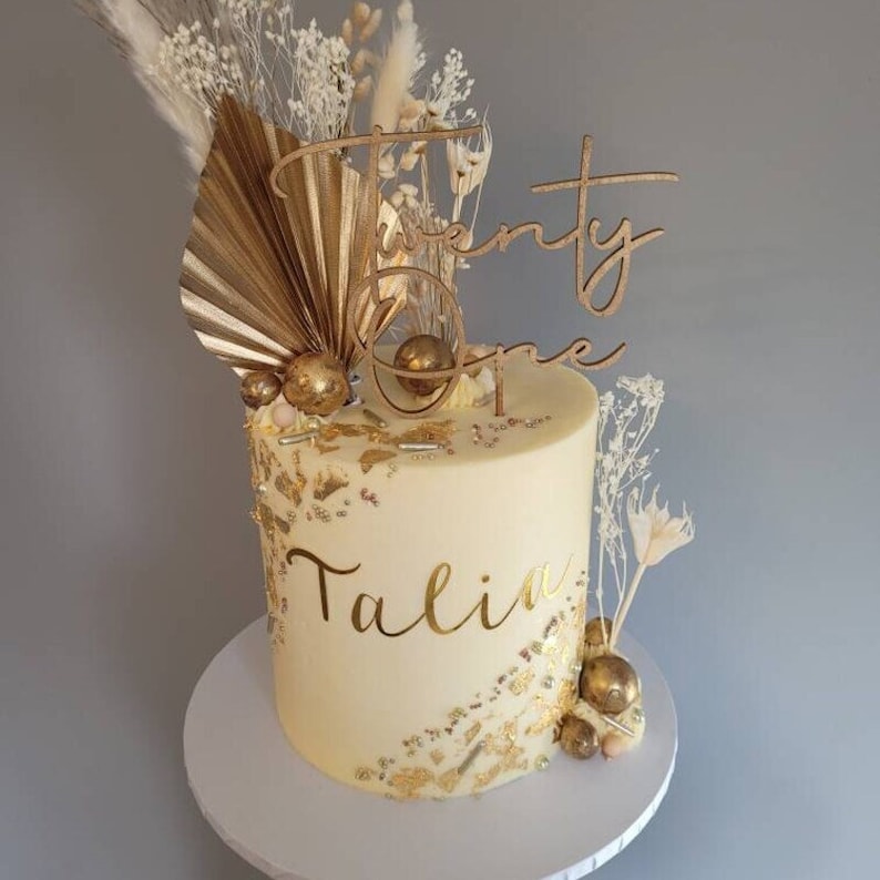 May include: A white cake decorated with gold accents, dried flowers, and a wooden topper that says "Twenty One". The cake is also decorated with the name "Talia" in gold lettering.
