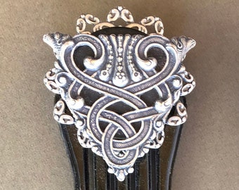 May include: A black plastic comb with a silver ornate design at the top. The design features a knotwork pattern with two small bird figures on either side.