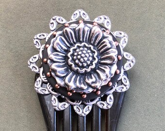 May include: A black tortoiseshell hair comb with a silver and copper flower design. The flower has a raised center and is surrounded by a decorative border.