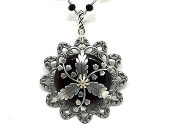 May include: A silver necklace with a black bead chain and a pendant featuring a dark red cabochon stone surrounded by a silver filigree design with leaves and flowers.