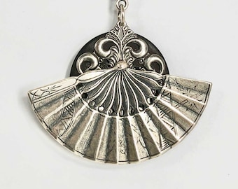 May include: A silver pendant necklace with a fan-shaped design and a circular pendant with intricate details. The necklace has a black chain.