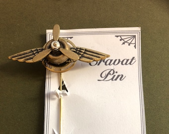 May include: A gold-toned metal airplane lapel pin with a propeller. The pin is attached to a gold-toned metal stick. The pin is on a white card with the text "Cravat Pin" and "Time Trinkets by Arynne Elfenbein" and the number "831.917.0124".