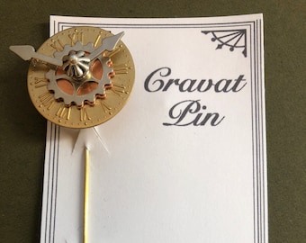 May include: A gold and silver clock face cravat pin with a gold pin. The pin is on a white card with the text "Cravat Pin", "TIME TRINKETS", "by Arynne Elfenbein", and "831.917.0124".