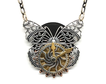 May include: A silver and gold butterfly necklace with a clock face in the center. The butterfly wings are detailed with filigree and the clock face has a gold gear design.
