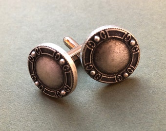 May include: A pair of silver cufflinks with a circular design. The cufflinks have a textured surface and a raised border.