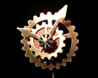 May include: A gold-toned metal pin with a gear-shaped design. The pin features a smaller gear in the center with a red and brown background. The pin is attached to a long, thin, gold-toned metal pin.