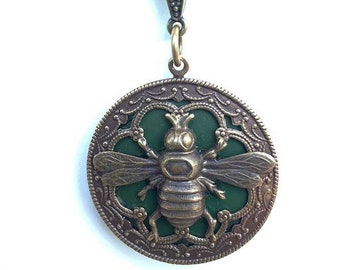 May include: A gold necklace with a round pendant featuring a green enamel bee with intricate details. The pendant is surrounded by a decorative filigree design.