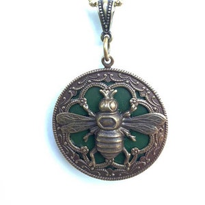 May include: A gold necklace with a round pendant featuring a green enamel bee with intricate details. The pendant is surrounded by a decorative filigree design.
