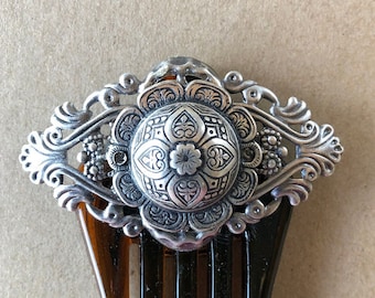 May include: A silver hair comb with an ornate, detailed design. The comb has a decorative, round, silver top with intricate floral patterns.