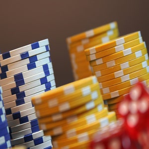 May include: Stacks of poker chips in various colors, including blue, white, and yellow. The chips are arranged in neat piles, with a few red dice in the foreground. The chips have a circular shape and are used for casino games.