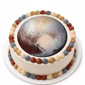 May include: A round cake with smooth white frosting featuring a large, circular edible image of the planet Pluto on top. The cake is decorated with a border of small, piped frosting swirls in alternating colors of tan, rust, and slate blue.