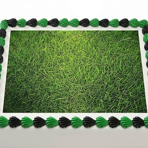 May include: A rectangular cake with a green grass design on top. The cake is decorated with green and black frosting along the edges. The cake is white.
