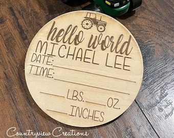 May include: A wooden round sign with the text "hello world MICHAEL LEE" and spaces for the baby's birth date, time, weight in pounds and ounces, and length in inches. A toy green tractor with a yellow trailer is in the background.