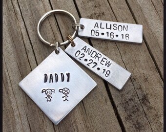May include: Silver keychain with three tags. The largest tag is a diamond shape with the word "Daddy" and stick figure drawings of a girl and boy. The other two tags are rectangular and have the names "Allison 05-16-16" and "Andrew 02-27-19". The text "Countryview Creations by Erika" is below the tags.