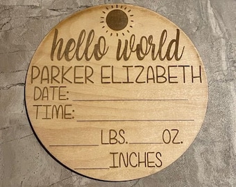 May include: A round wooden sign with the text "hello world" and a sun graphic. The sign also includes the text "PARKER ELIZABETH" and spaces to fill in the date, time, weight in pounds and ounces, and length in inches. The sign is made by Countryview Creations.