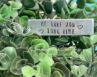 May include: Silver keychain with a heart and the text "ME LOVE YOU LONG TIME" on a silver rectangle. The keychain is on a green plant.