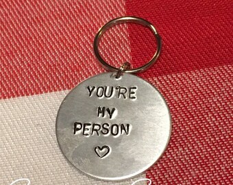 May include: Silver keychain with a round charm that says "You're My Person" with a small heart symbol. The keychain is on a red and white checkered background.