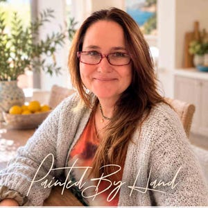 May include: A person with long brown hair wearing red-rimmed glasses, a grey textured cardigan, and an orange patterned top. The image features the text "Painted By Hand" in a cursive script and "Kate Shephard Art" in a dark serif font below.