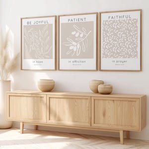 May include: Three framed wall art prints with light beige backgrounds and white line art illustrations. The prints feature the text BE JOYFUL in hope, PATIENT in affliction, and FAITHFUL in prayer, each with a light wood frame and white matting.