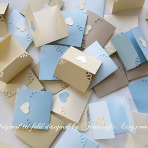 May include: A collection of folded cards with a heart cut-out design. The cards are in shades of blue and beige. The text "Original trifold designed by Trisicrafts. Etsy.com" is visible at the bottom of the image.